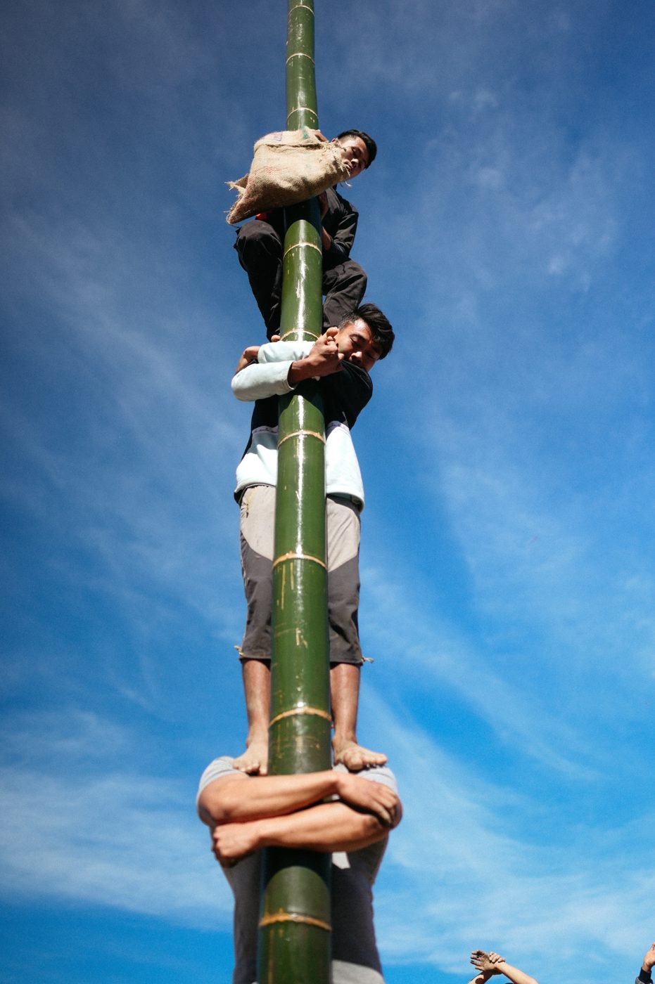 Three young men climbing a tall bamboo pole against a blue sky, one standing on another's shoulders