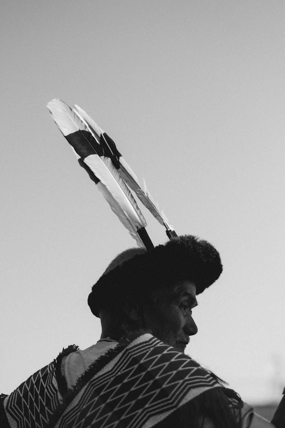 An elder in a fur headdress with two long feathers, seen in profile in black and white