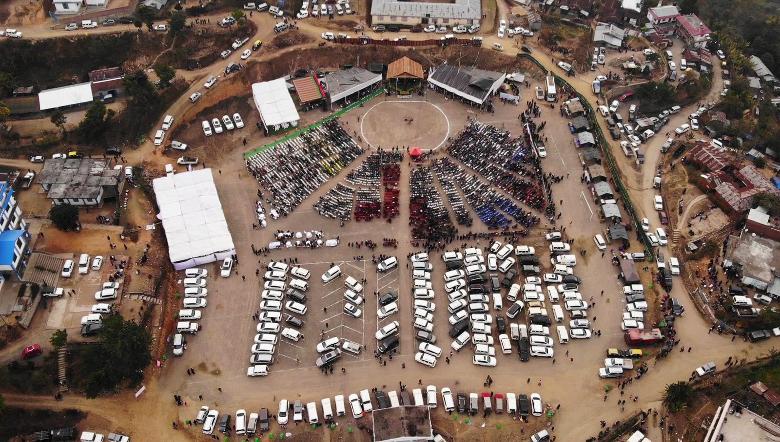 Aerial view of a crowd seated in blocks around a festival ground in Tseminyu, ringed by parked vehicles