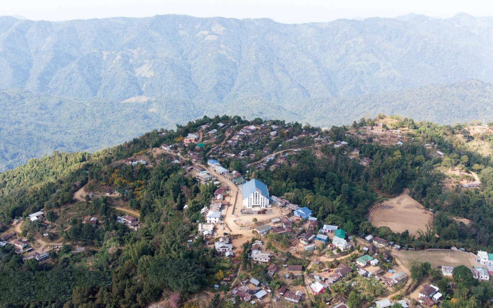 Aerial view of Sendenyu village on a forested ridge, its church and football ground in the centre, with ranges receding into haze